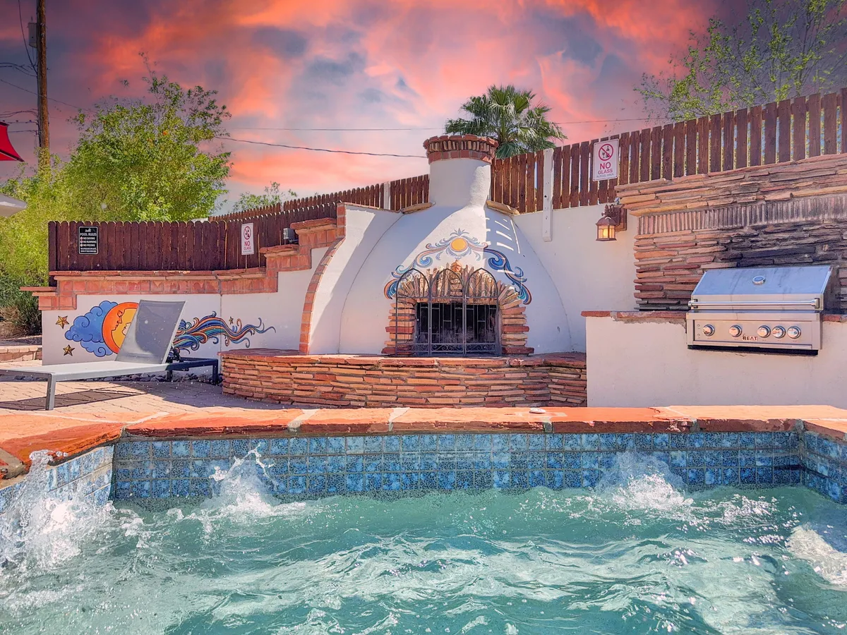 A welcoming outdoor kitchen is featured next to a modern pool, complete with an inviting dining area and a striking sun and moon mural on the chimney. The scene is set for al fresco dining and socializing by the poolside, with soft evening lighting and a backdrop of the Arizona sky.