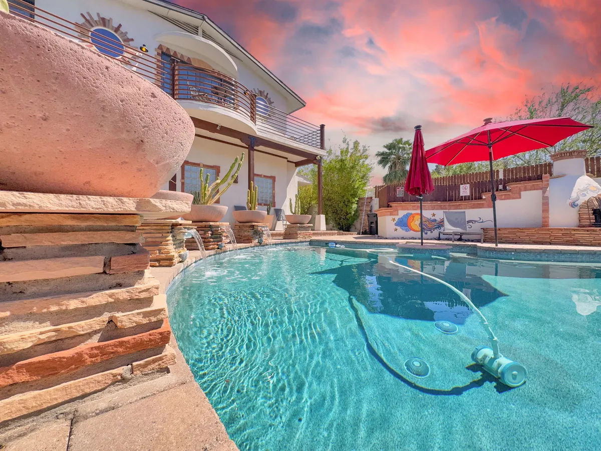 A tranquil backyard area featuring a swimming pool, red umbrella, stone walls, the house's facade, and potted plants. The scene is quiet and serene, suggesting a private retreat for both humans and their pets to relax and soak up the sun.