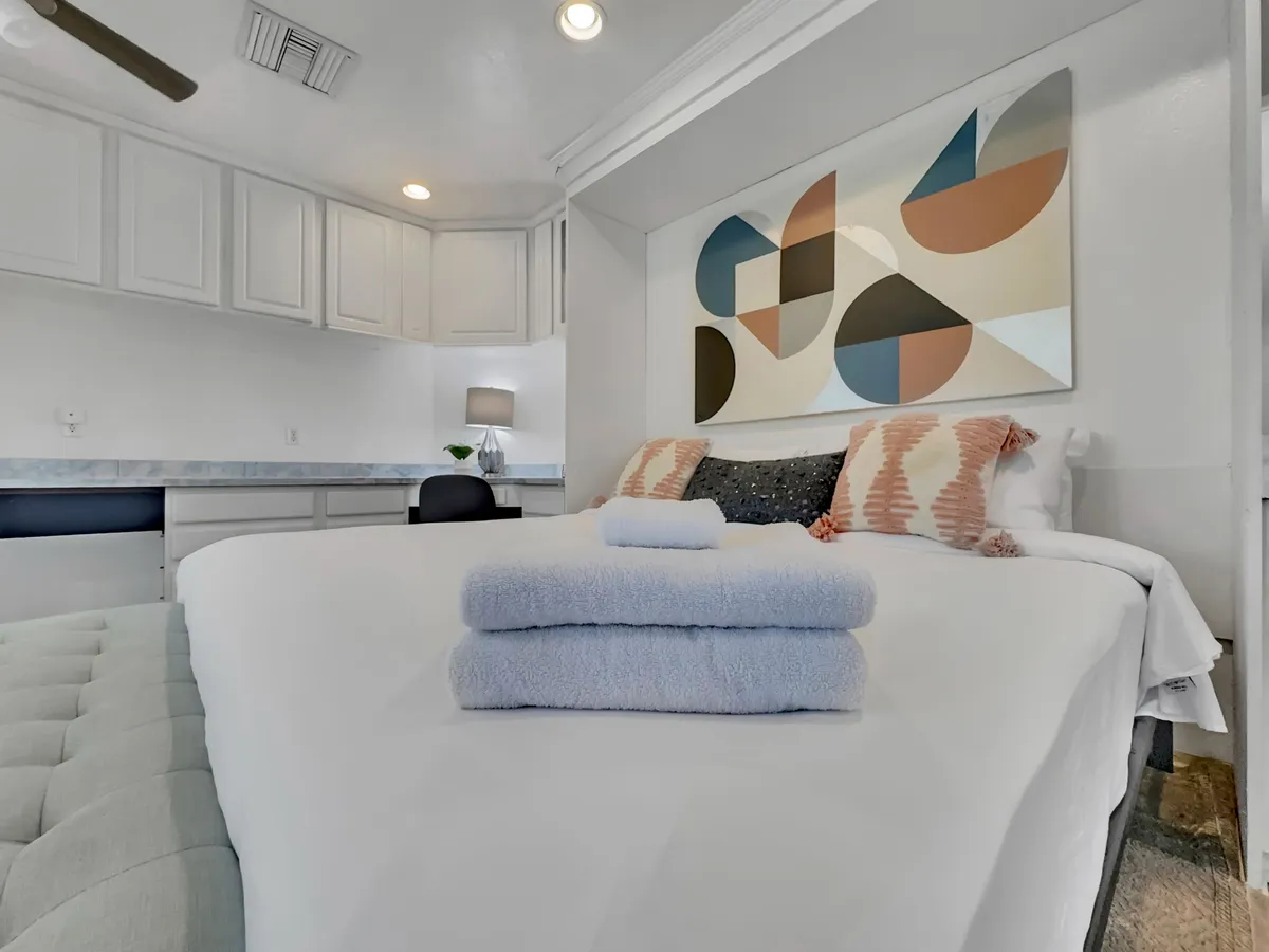 A serene bedroom with a neatly made bed, decorative pillows, abstract wall art, and folded towels, bathed in natural sunlight. The room’s soft color palette and uncluttered layout evoke a restful, cozy atmosphere perfect for unwinding.
