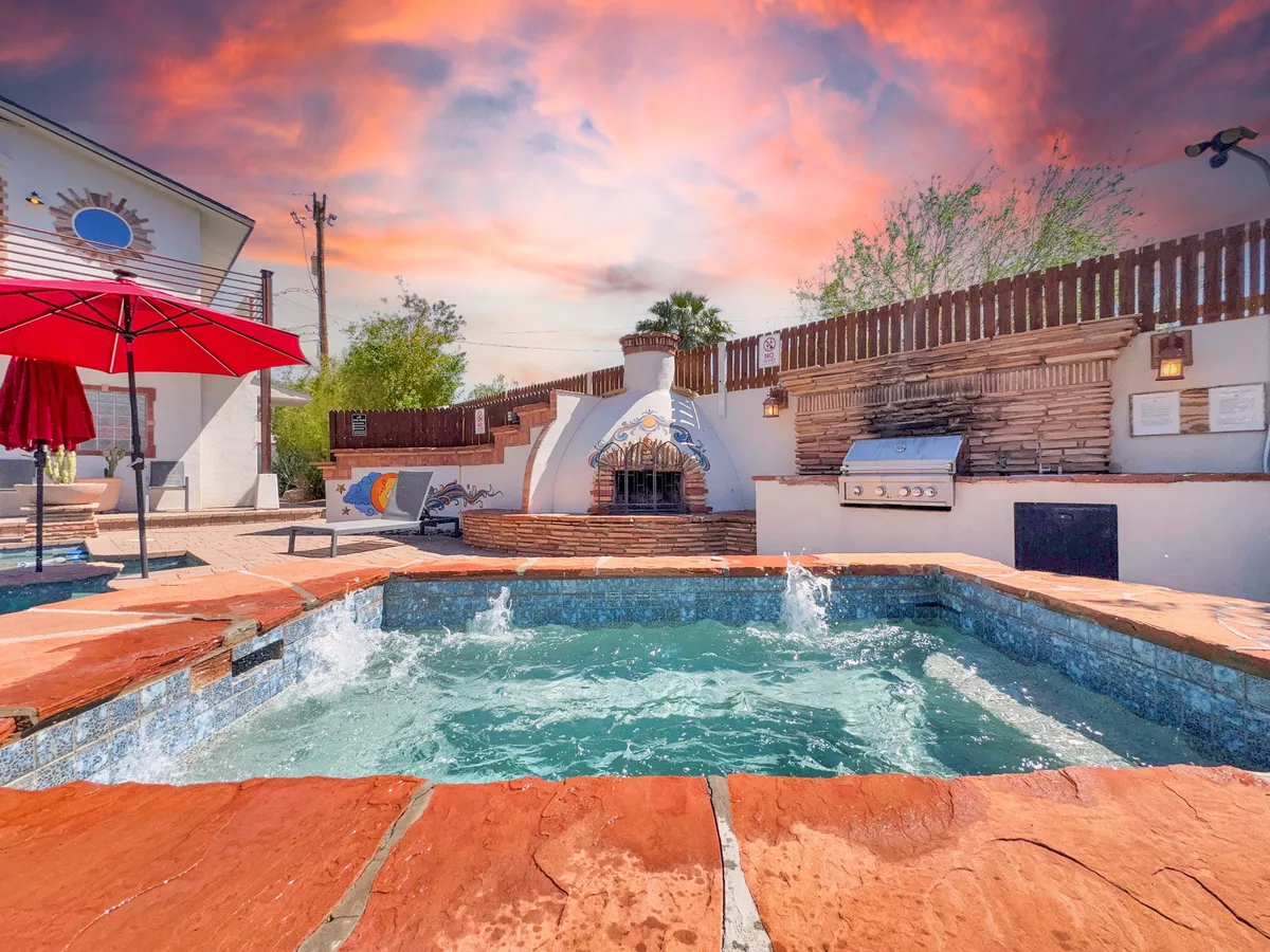 A cozy evening view of the outdoor living space, spotlighting the pool, an outdoor fireplace, grill area, and a bold red umbrella. The scene is set for sunset dinners, warm gatherings around the fire, and fun poolside evenings.