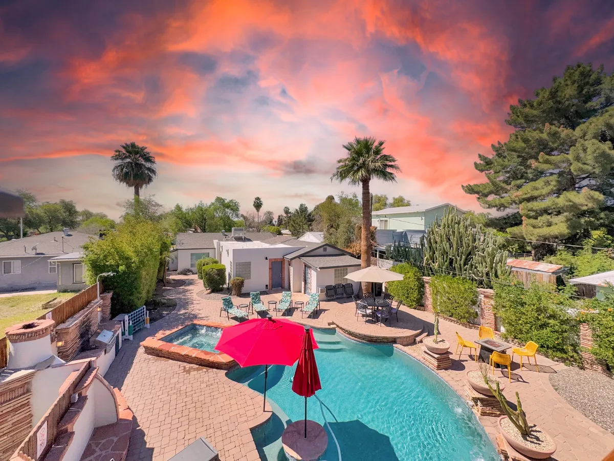 A broad view of the backyard retreat, highlighting the swimming pool, vibrant red umbrellas, multiple lounge chairs, and iconic desert landscaping with palm trees and cacti. The scene exudes a relaxed, sun-drenched ambiance ideal for family fun or quiet moments by the water.