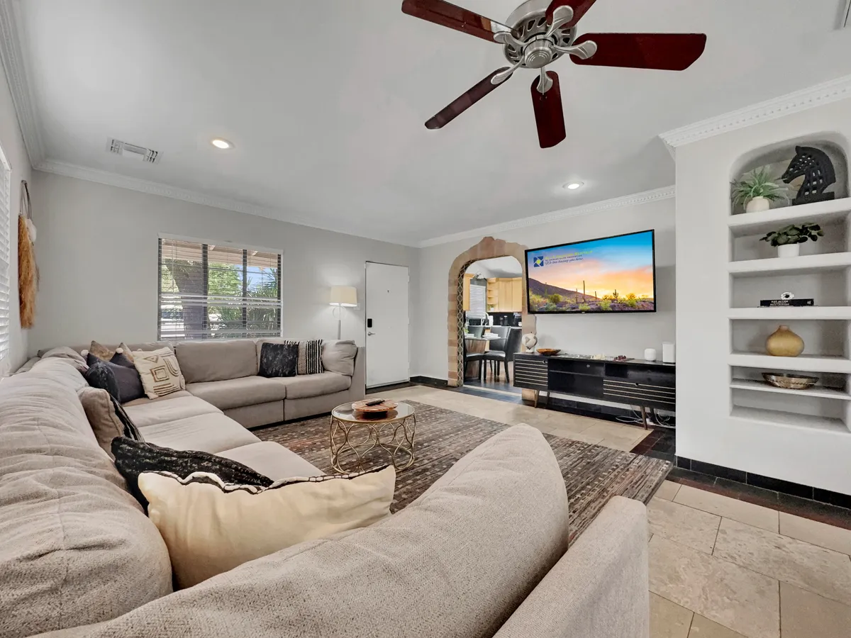 A spacious open-concept living area featuring a large sectional sofa, decorative coffee table, television mounted on the wall, and an arched entrance leading to the kitchen. The layout is welcoming and designed for family gatherings, conversations, and relaxed entertainment.