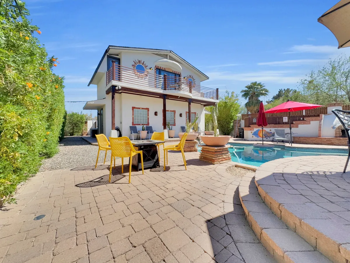 A vibrant exterior view of the home's swimming pool area, featuring yellow chairs, a patio table, and the house facade. This lively setting is framed by clear skies and lush landscaping, inviting groups to gather and celebrate.