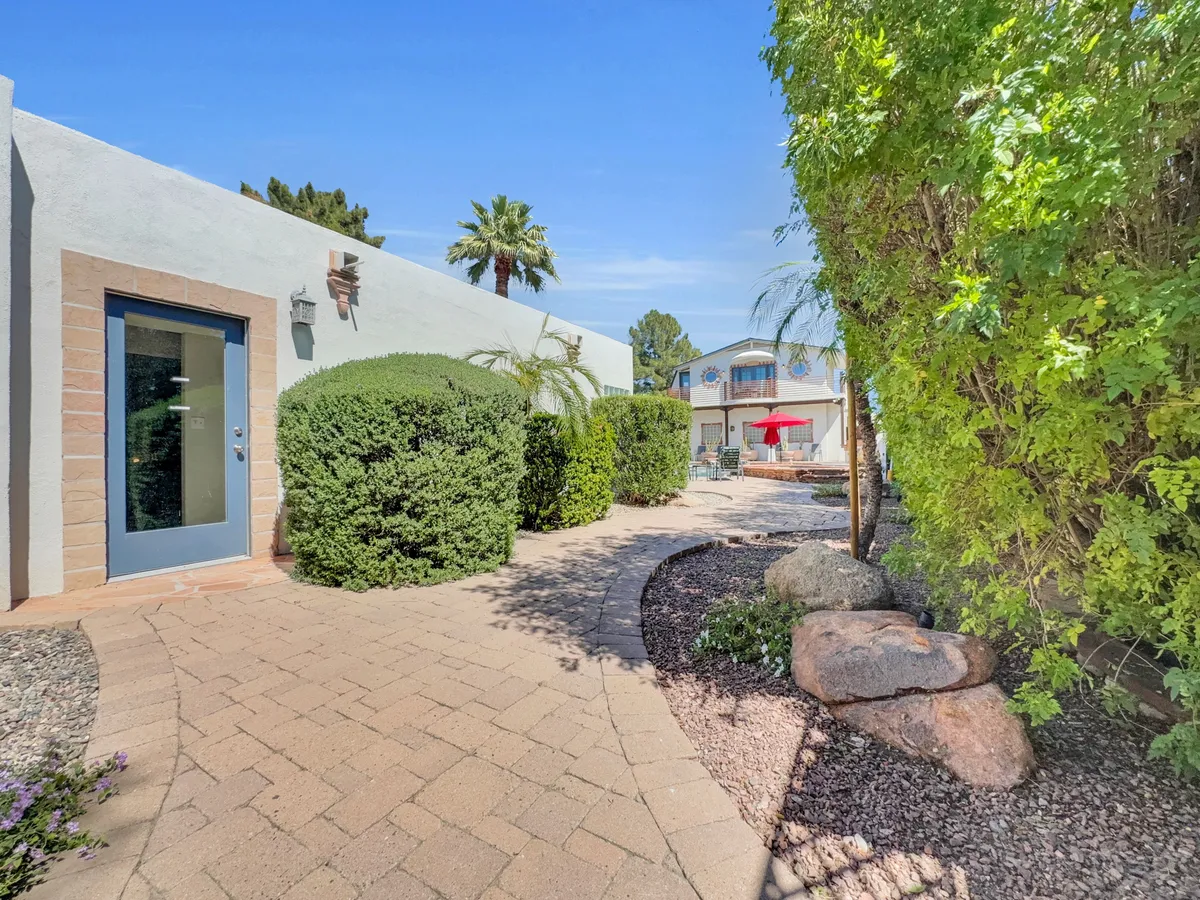 This exterior shot features a welcoming pathway leading through a lush garden to the house, accented by palm trees and a glimpse of a pool area with a vibrant red patio umbrella. The tranquil landscape and open space evoke a sense of arrival and outdoor enjoyment.