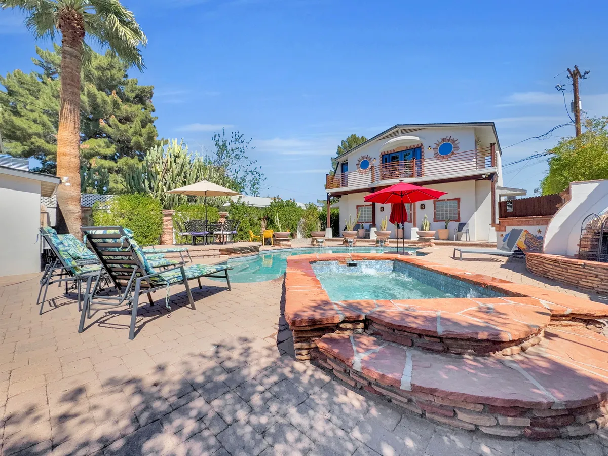 This image captures a vibrant backyard scene with a sparkling pool, a bubbling hot tub, and a cheerful red umbrella surrounded by palm trees and the house. Lounge chairs invite guests to relax in the Arizona sun, and the lush landscaping frames the scene in tranquility and privacy.