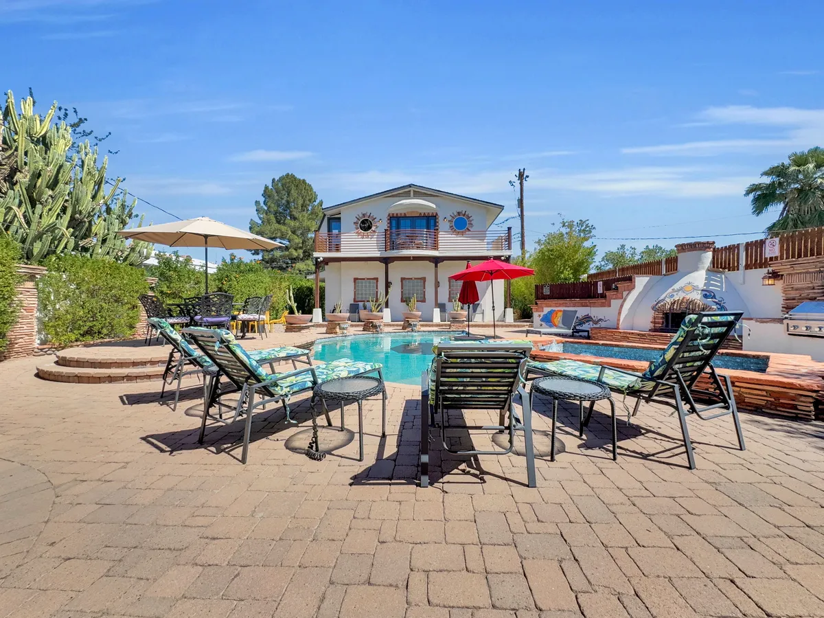 This exterior image showcases a sunlit swimming pool bordered by lounge chairs, a red umbrella, potted plants, and the inviting facade of the home. The setting exudes leisure, with ample space for groups to gather and relax poolside or dine outdoors together.